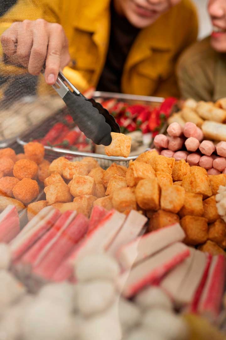 Tightly cropped image of people serving themselves food from a buffet of cheese cubes, sausages, slices of meat, and other finger foods.