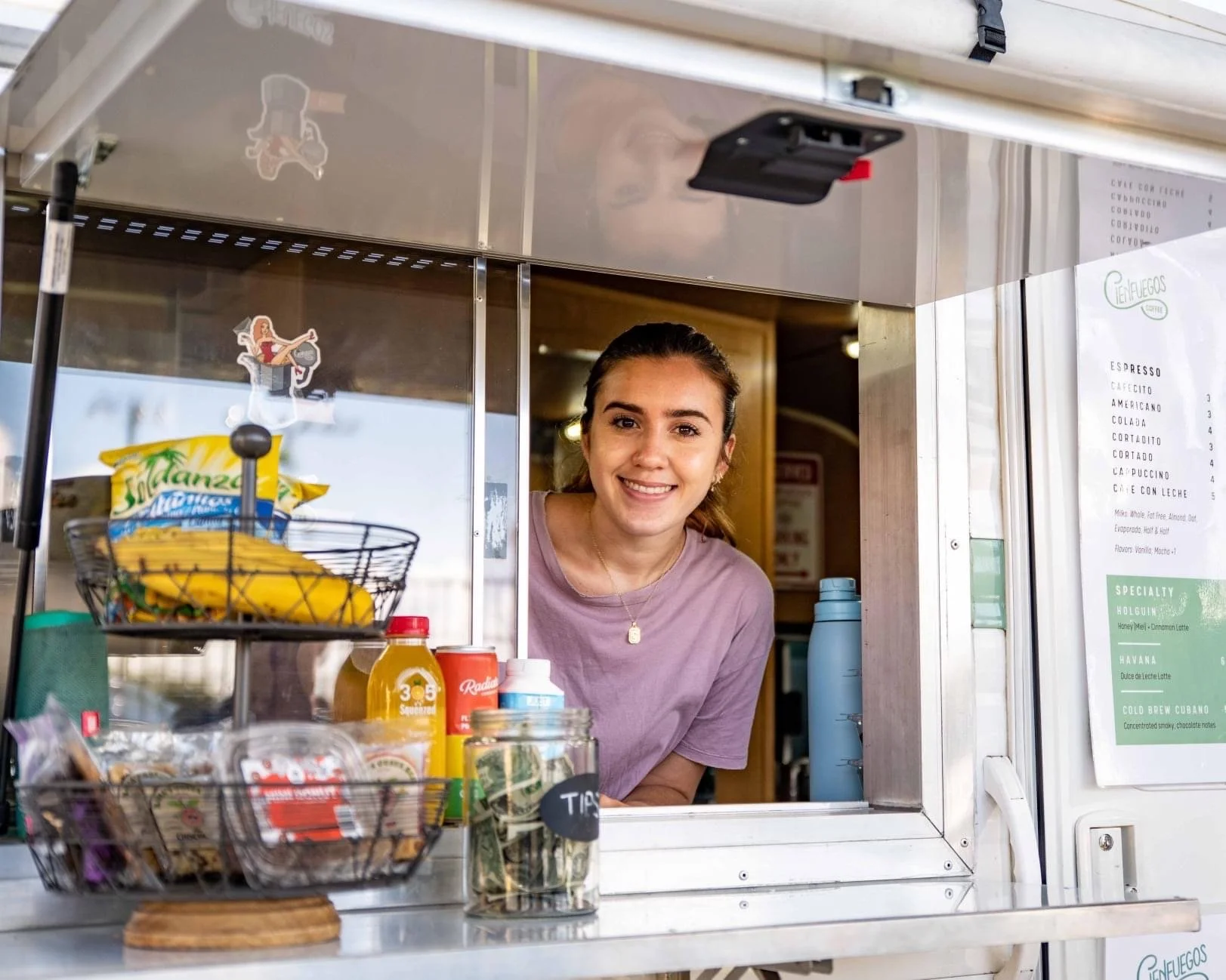 Smiling barista leaning out of a food truck service window, with a menu board, beverage bottles, and a tip jar visible on the counter inside the truck.