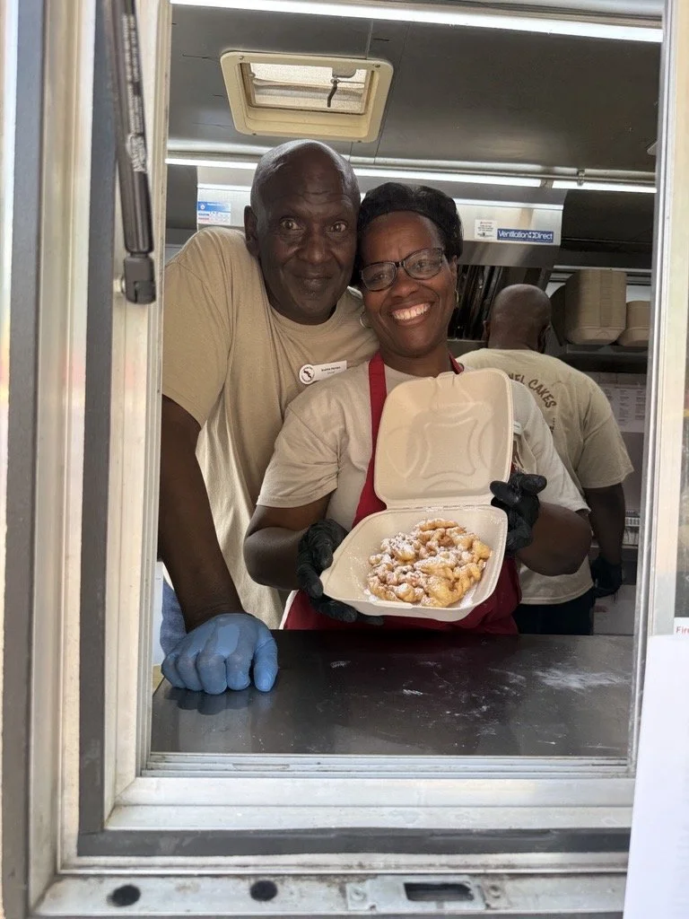 Smiling couple inside a food truck, with the woman holding a takeout container filled with powdered-sugar–topped funnel cake as the man leans in beside her.