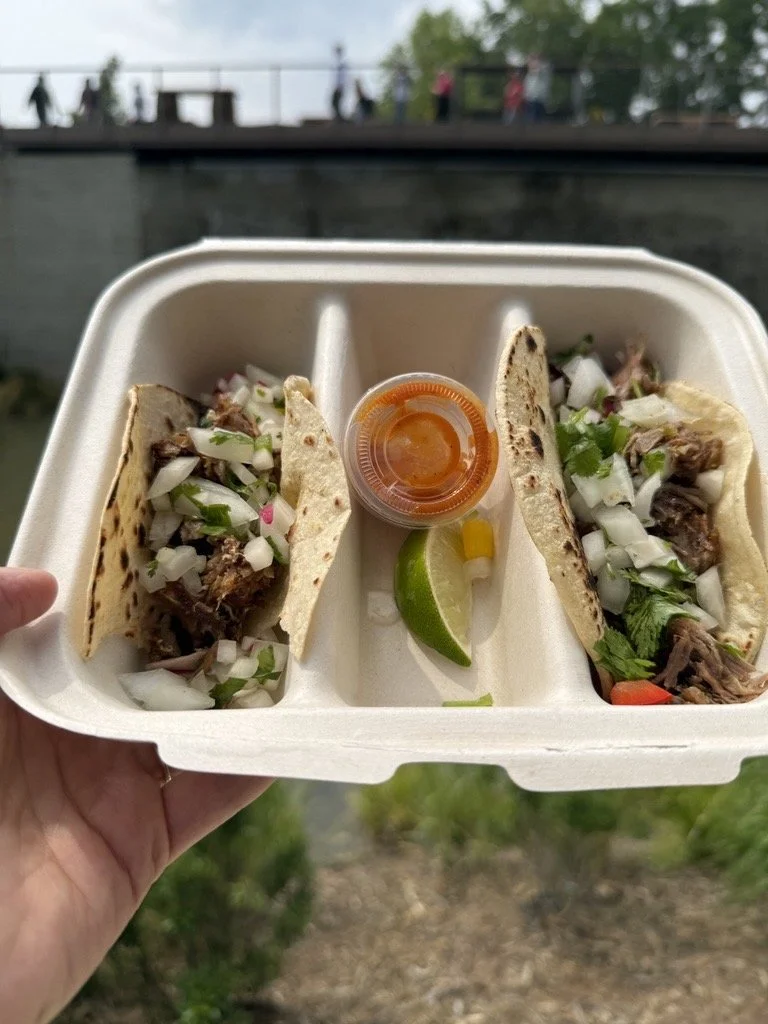Hand holding a compostable takeout container with two tacos filled with shredded meat, diced onions, and cilantro, accompanied by a small cup of red sauce and a lime wedge, with an outdoor setting blurred in the background.
