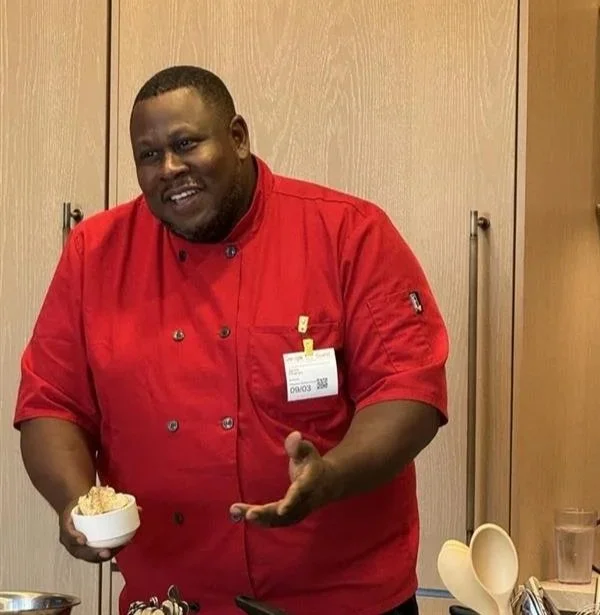 Chef wearing a red coat smiling and gesturing while holding a small bowl of food in a kitchen or demonstration setting.