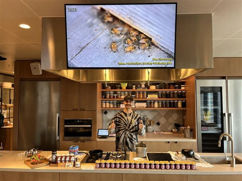 Person standing behind a kitchen demonstration counter giving a thumbs-up, with jars and cooking ingredients arranged on the counter and a large screen overhead displaying a close-up image of bees.
