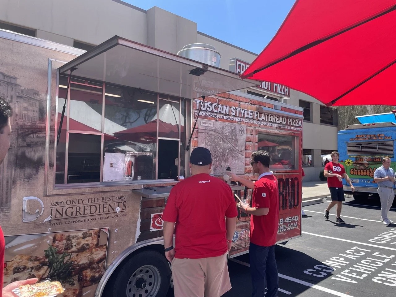 Two people in red shirts standing at the window of a Tuscan-style flatbread pizza food truck, ordering food under bright red umbrellas on a sunny day.