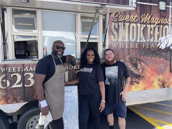 Three people standing and smiling in front of the Sweet Magnolia Smokehouse food truck, with one wearing an apron and the others dressed casually.