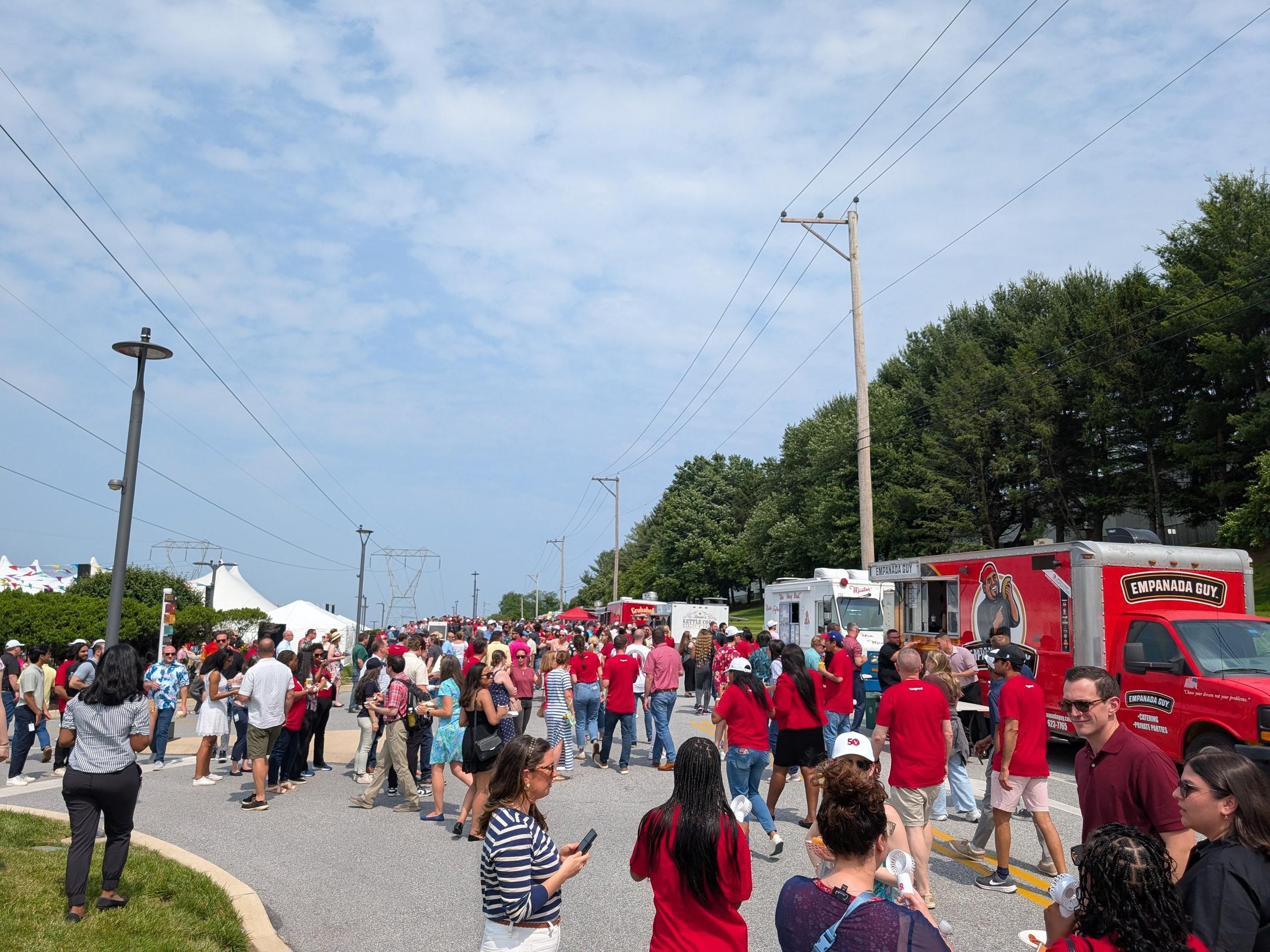 Large outdoor festival with crowds of people walking between rows of food trucks on a sunny day, with power lines and trees lining the road.