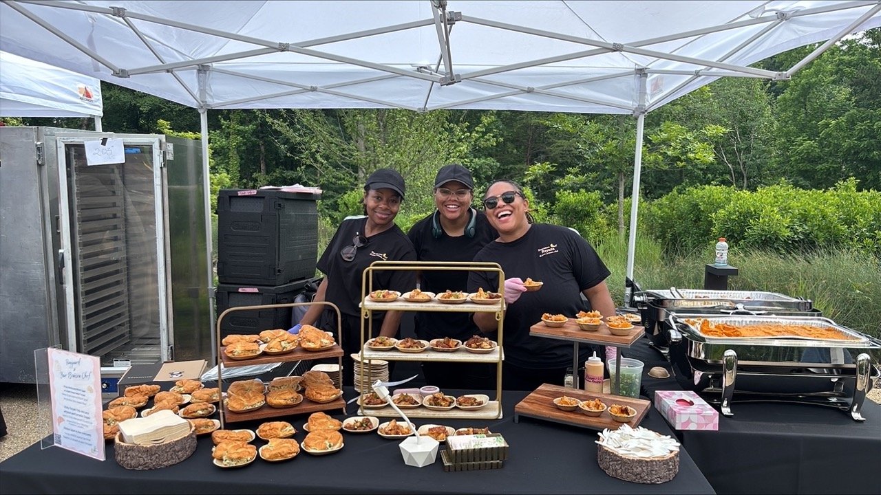 Three smiling women standing behind an outdoor food display under a canopy, presenting small plated dishes arranged on tiered stands and wooden trays.