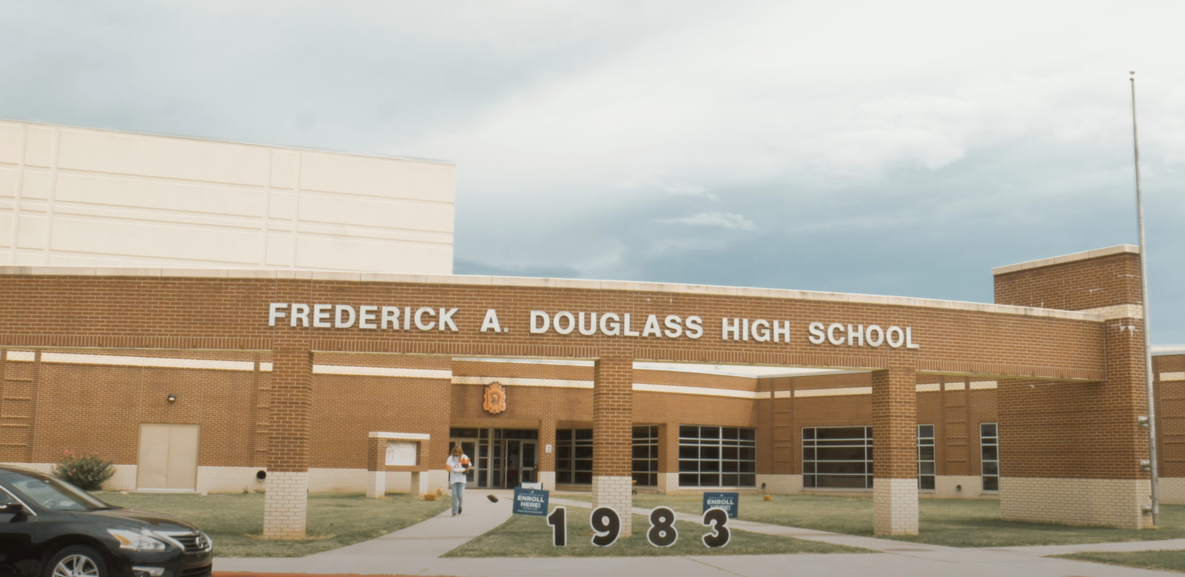 The exterior of Frederick A. Douglass High School building with brick walls and a sign displaying the year 1983. A person is walking on the sidewalk towards the entrance, and parked cars are visible in the foreground.