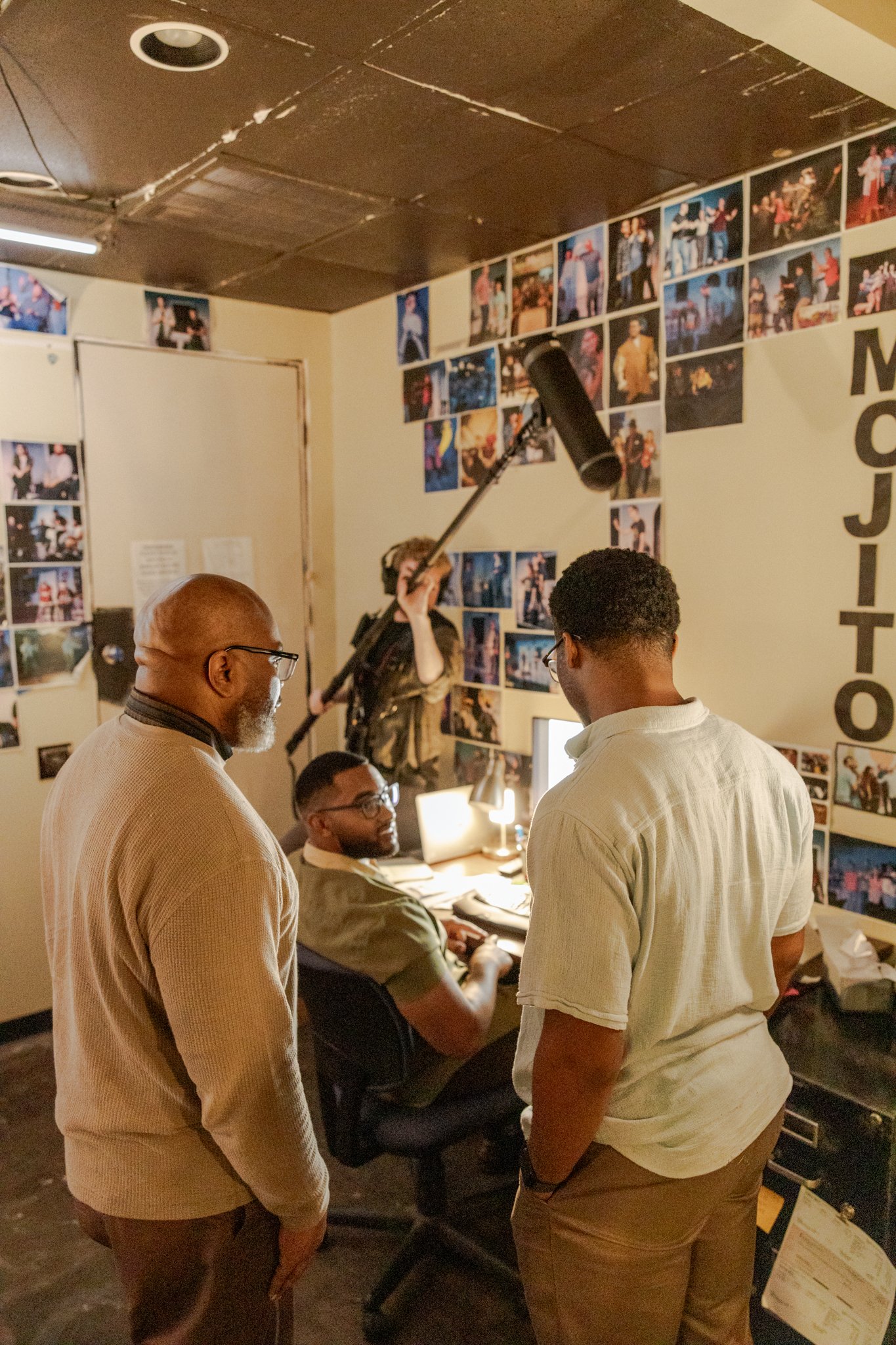 Titus Rhodes with a film or video production crew filming in an office. Four people are present, with one person holding a boom microphone overhead, all surrounded by wall decorations with numerous photos.