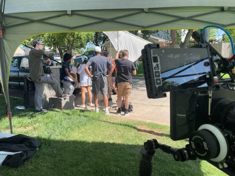 Titus Rhodes with a film crew shooting a scene outdoors under a green canopy, with equipment and people gathered around, adjacent to a street with a parked black pickup truck and trees.