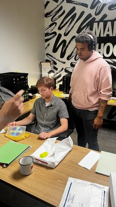 A young man with light skin, reddish hair, and a short beard sitting at a desk with a person pointing at something. The young man is wearing a grey short-sleeved shirt, and the person standing next to him has darker skin, short curly hair, headphones, a pink hoodie, and a tattoo on their arm. The desk has notebooks, a mug, a plastic bag, and a CD. The background includes a wall with black and white graffiti-style writing.