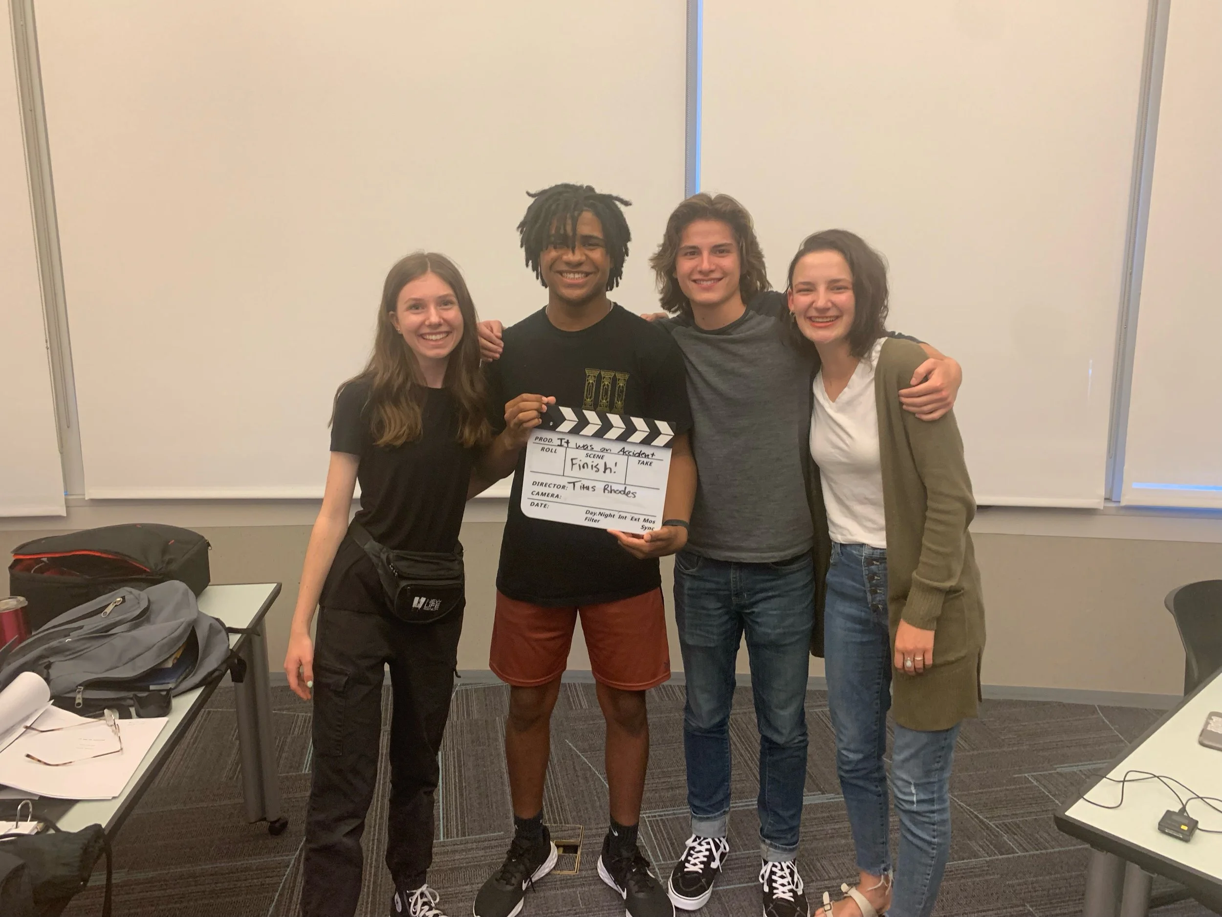 Group of four young adults standing together in a classroom, one holding a director's clapboard.