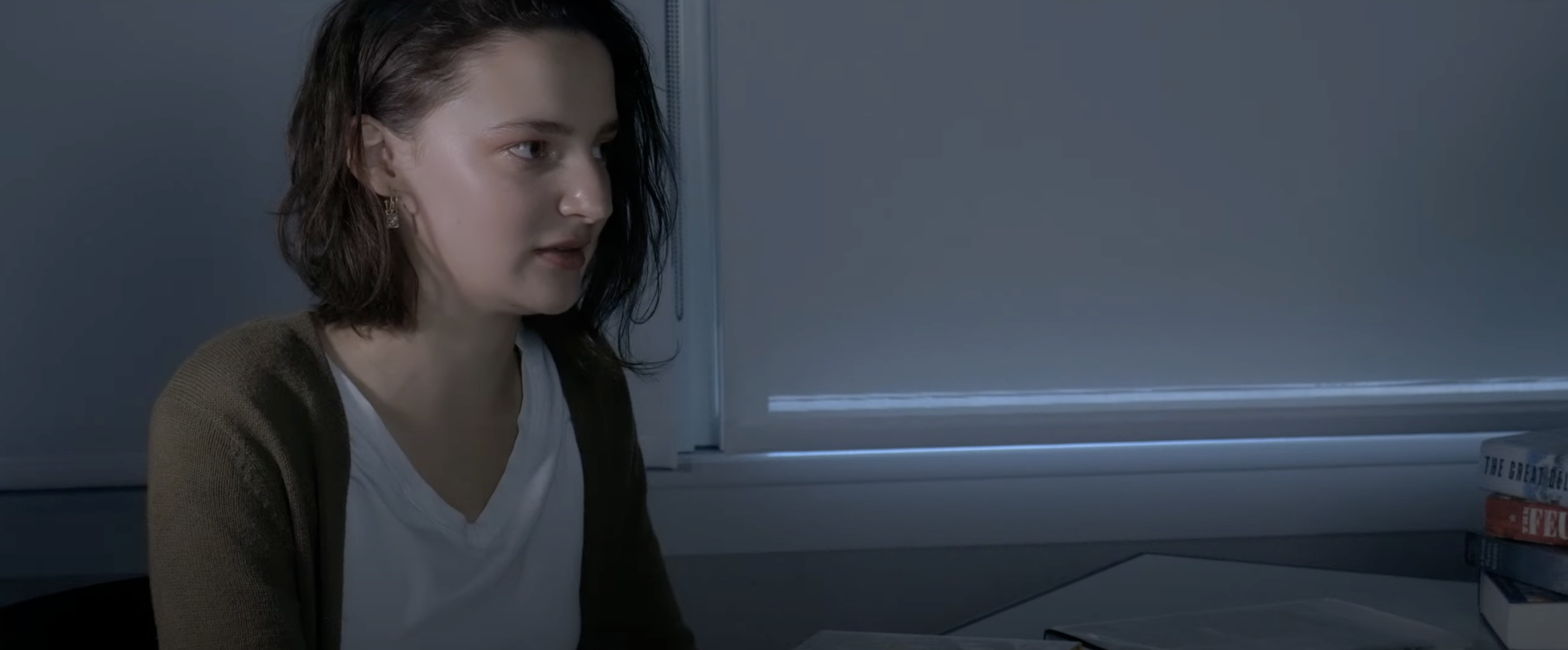 A woman with shoulder-length dark hair and earrings sitting at a desk in front of a window, with a stack of books nearby.
