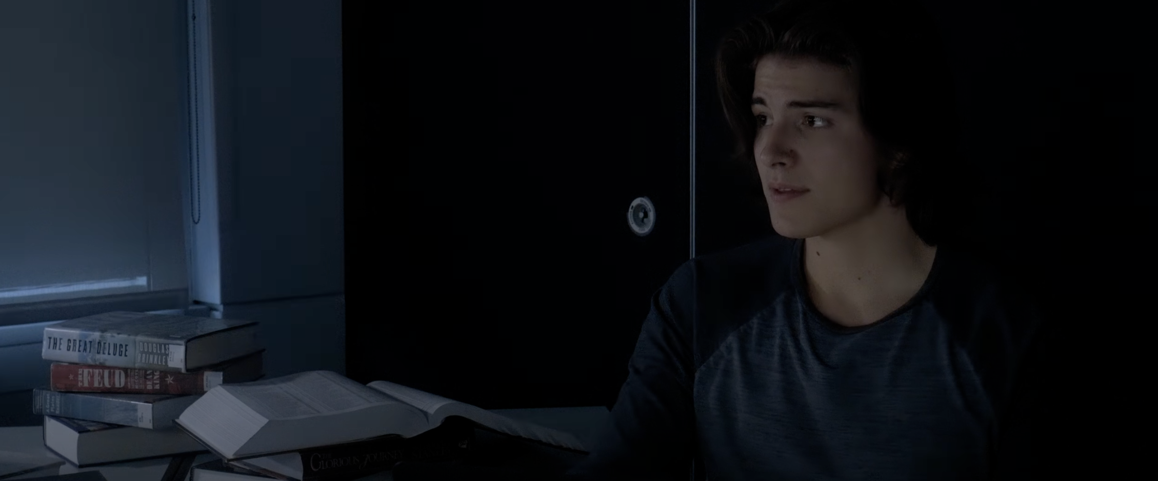 A young man with dark hair sitting at a desk with open books, wearing a dark shirt, in a dimly lit room.