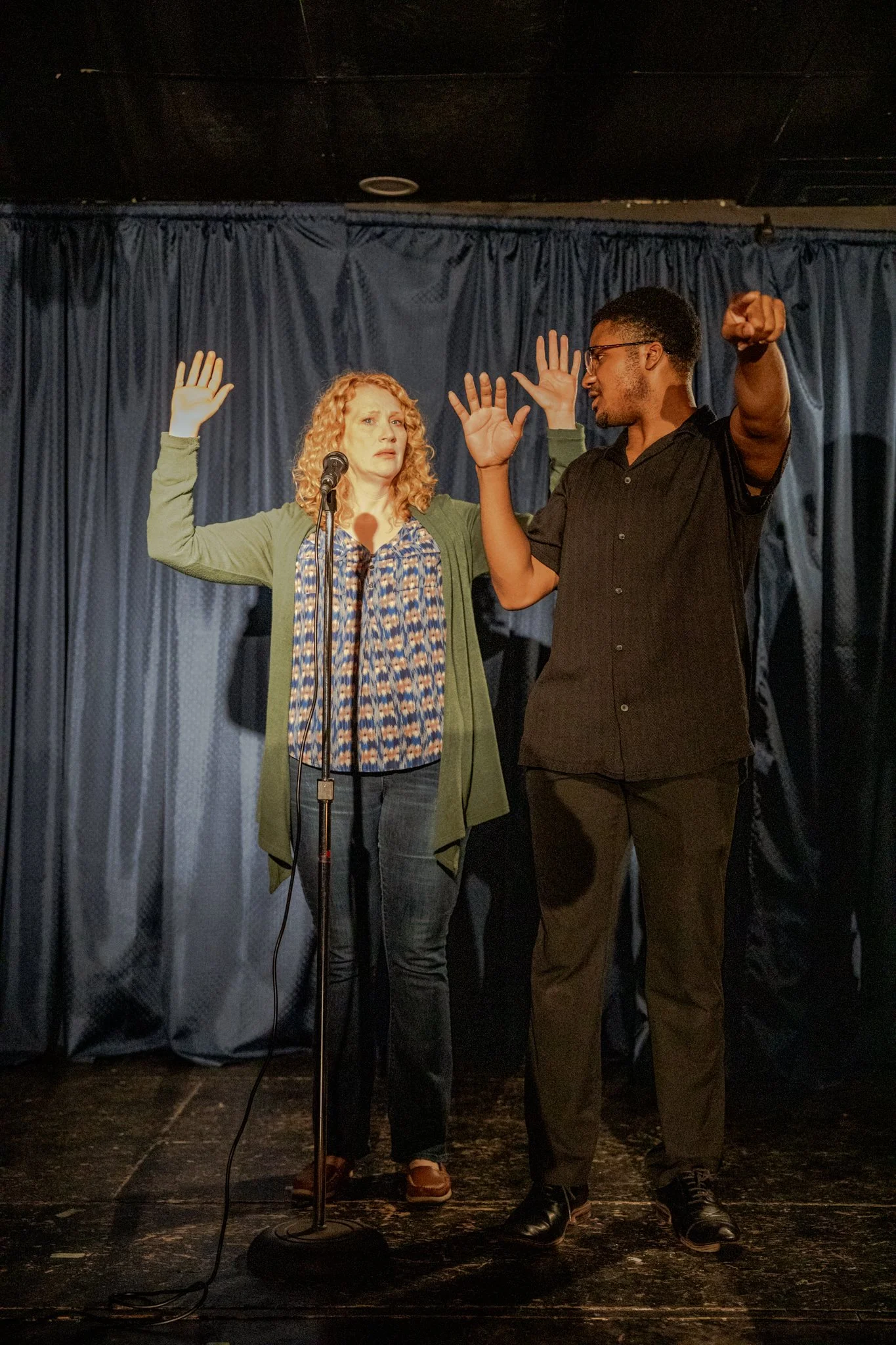 Titus Rhodes and Renee Krapff performing on stage, one woman with curly hair and a microphone, and one man with glasses, raising their hands, against a dark curtain backdrop.