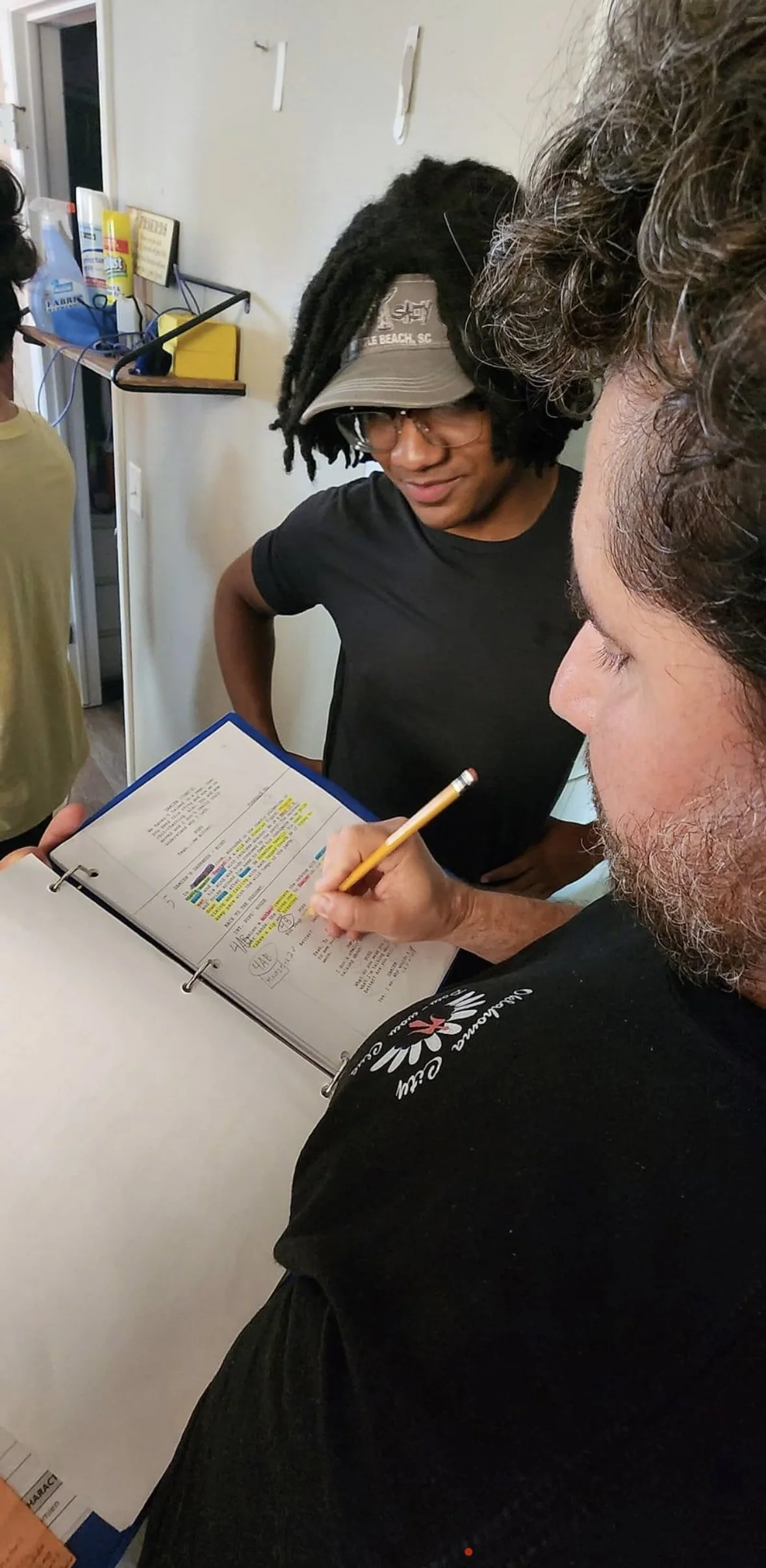 Two men discuss notes in a binder, one with dreadlocks and wearing glasses and a hat, the other with curly hair and a beard. Titus Rhodes looks down at the binder while the man with curly hair writes in it, both in casual black t-shirts.
