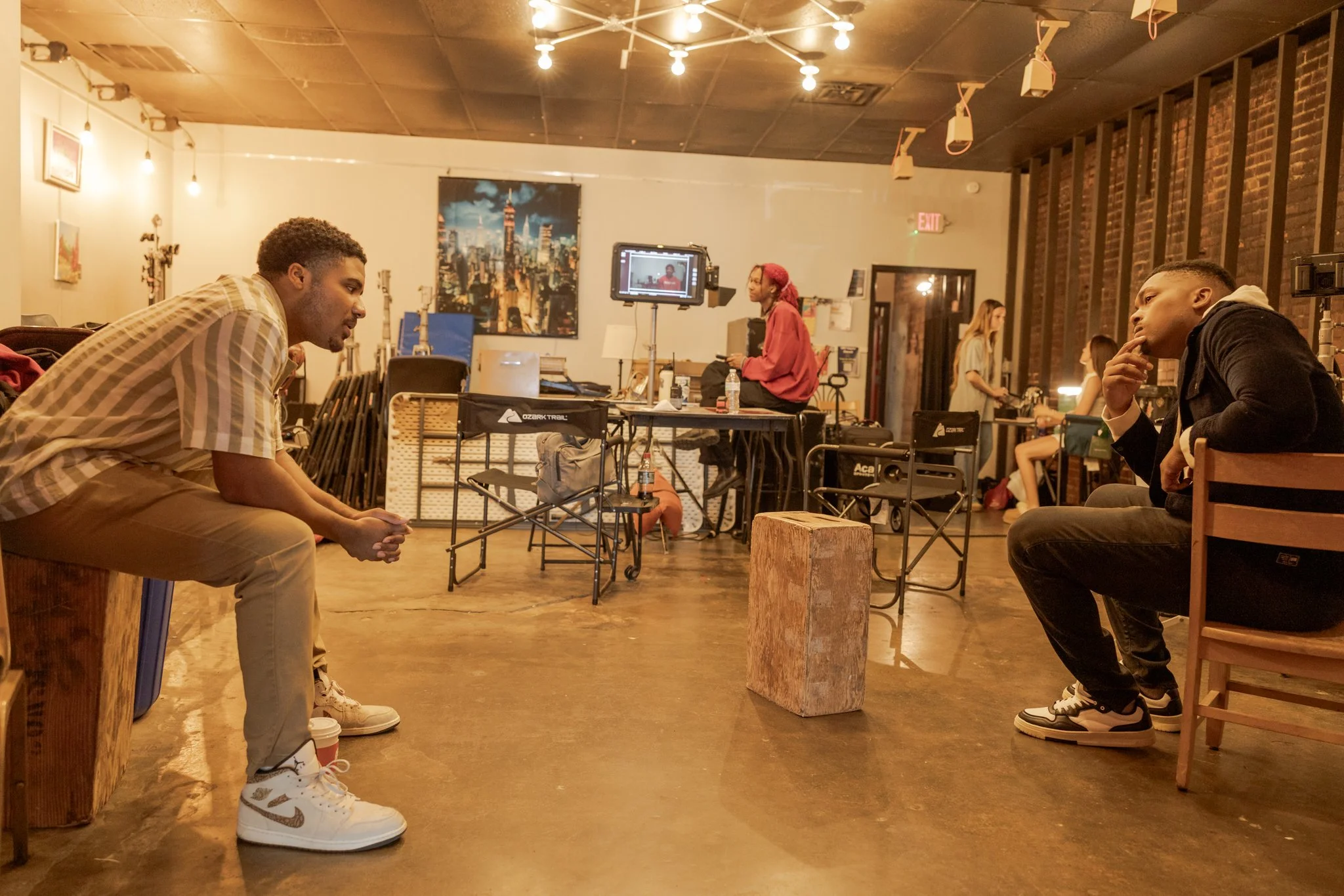 Titus Rhodes and Daven Joseph sitting and facing each other in conversation in a studio, with a woman working in the background, various equipment, and artwork on the walls.