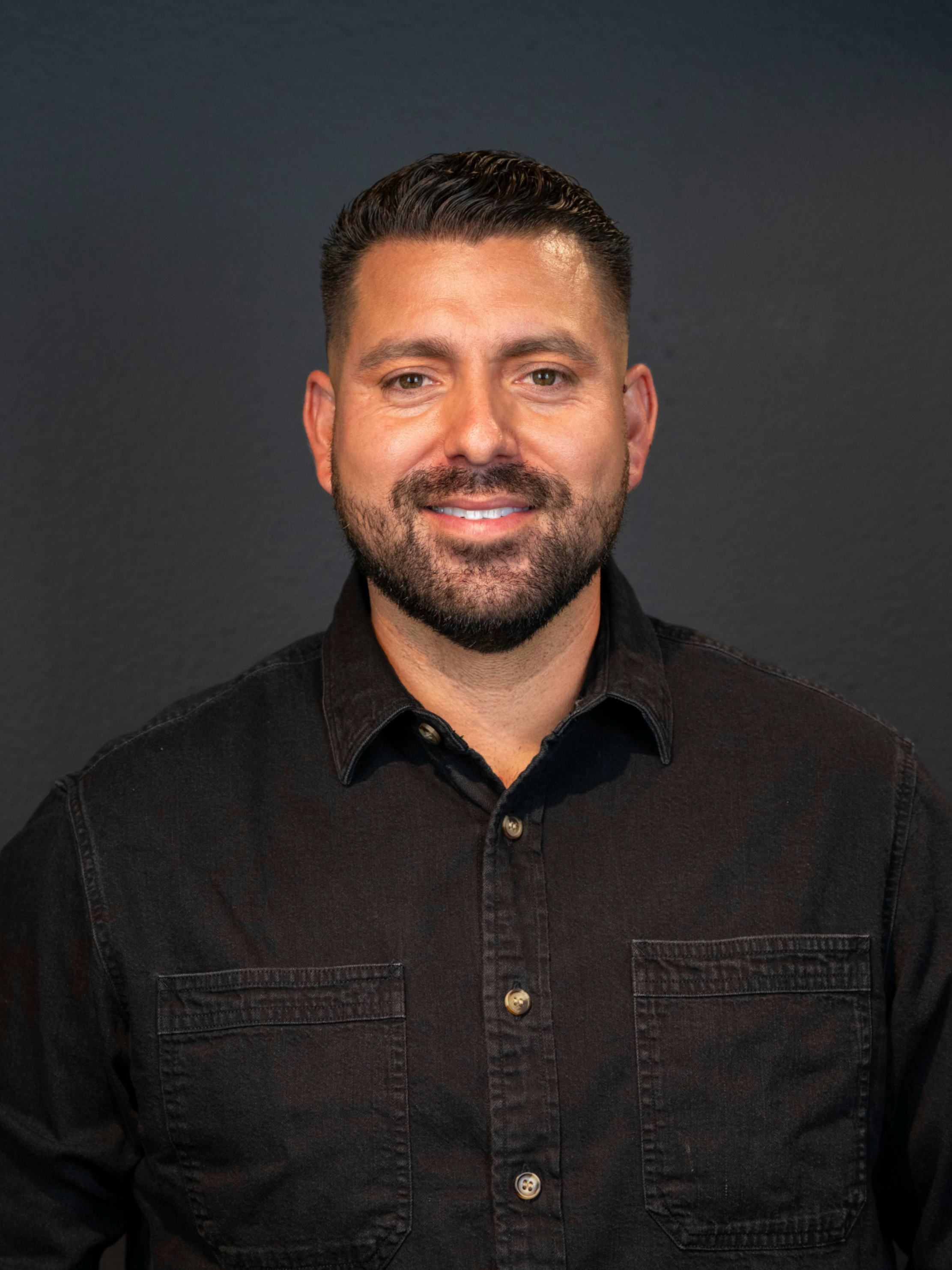 Head and shoulders portrait of a smiling man with dark hair, beard, and mustache, wearing a black collared shirt against a dark background.