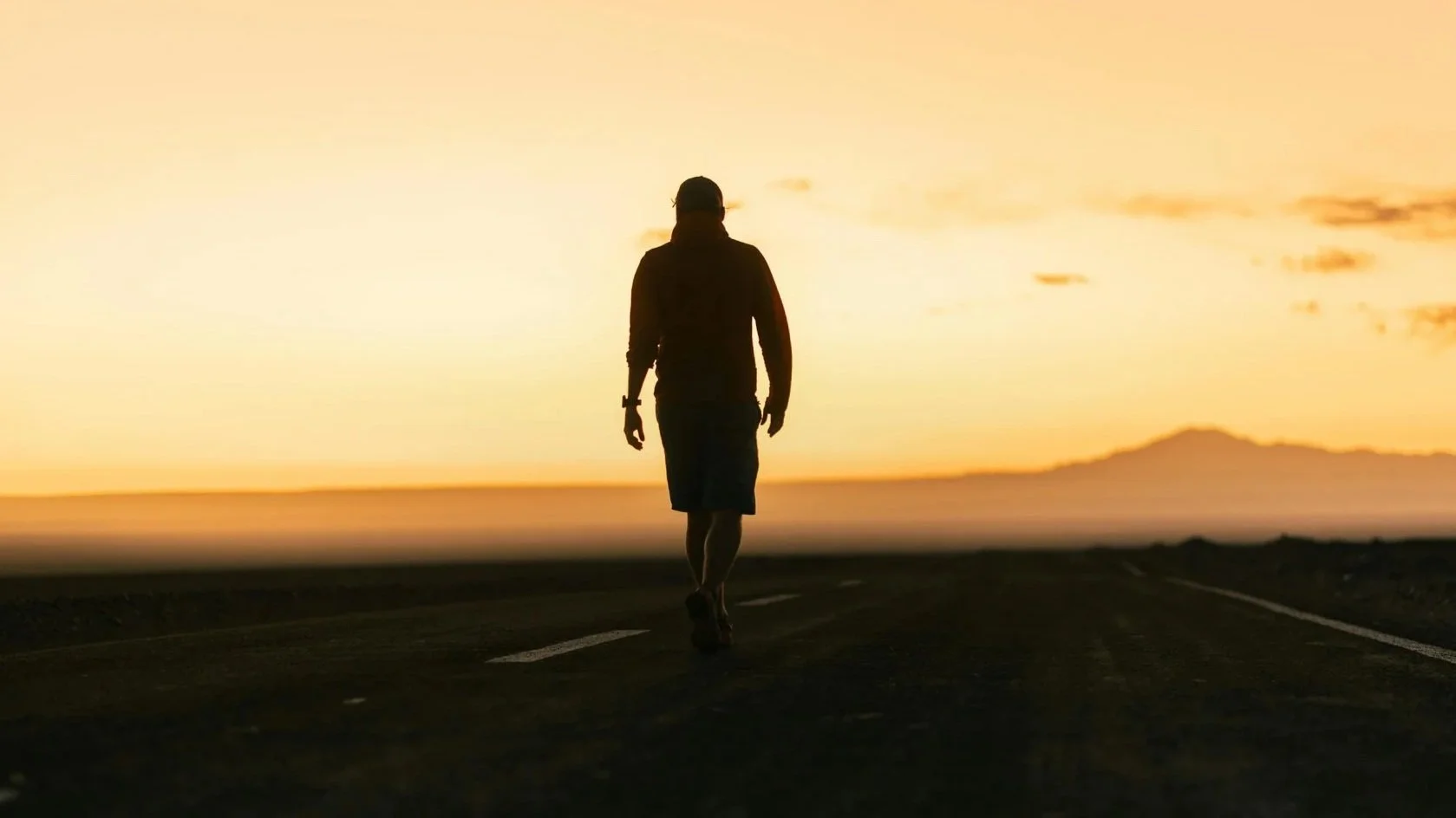 A person walking down a deserted road at sunset, with mountains in the distance and the sky painted in warm tones.
