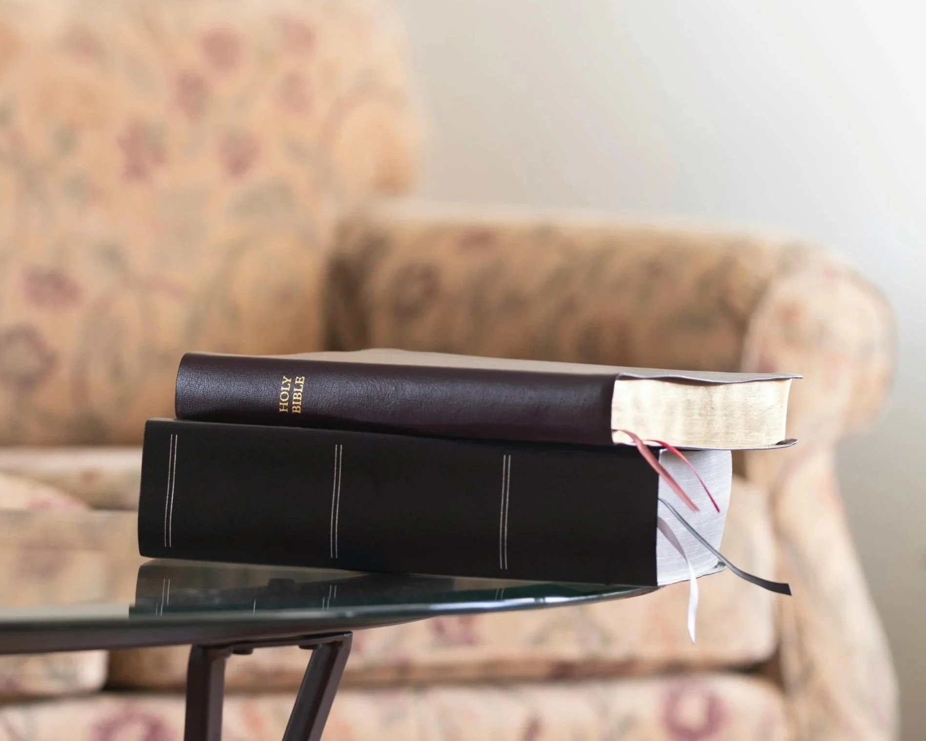 Three stacked books on a glass table with a couch in the background. The top book is a brown Bible.