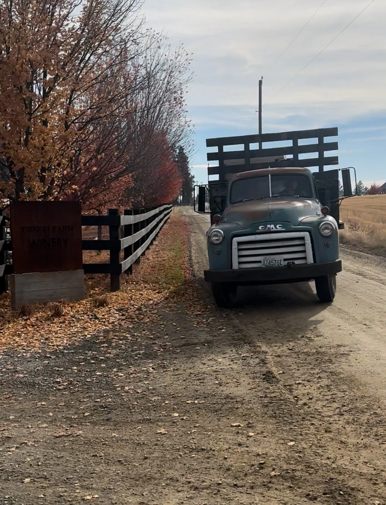 A vintage GMC truck parked on a dirt road near a black wooden fence. Trees with autumn leaves and a sign indicating a winery are visible on the left side, under a partly cloudy sky.