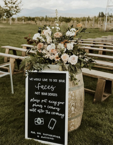 Wine barrel with floral display on top with chalkboard sign leaning against the front with ceremony benches behind.