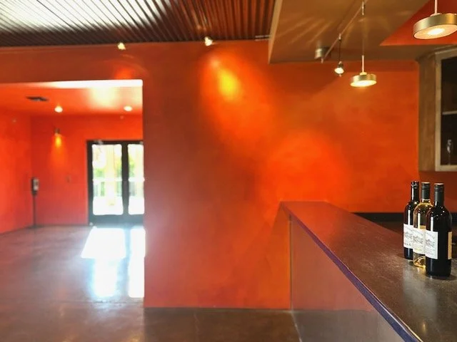 Interior of a wine tasting room with orange walls, a door with glass panels, a bar counter with three wine bottles, and modern light fixtures.