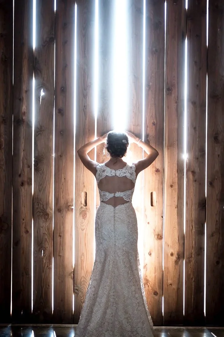 A woman in a white lace wedding dress standing in front of a tall barn door with light coming through gaps.