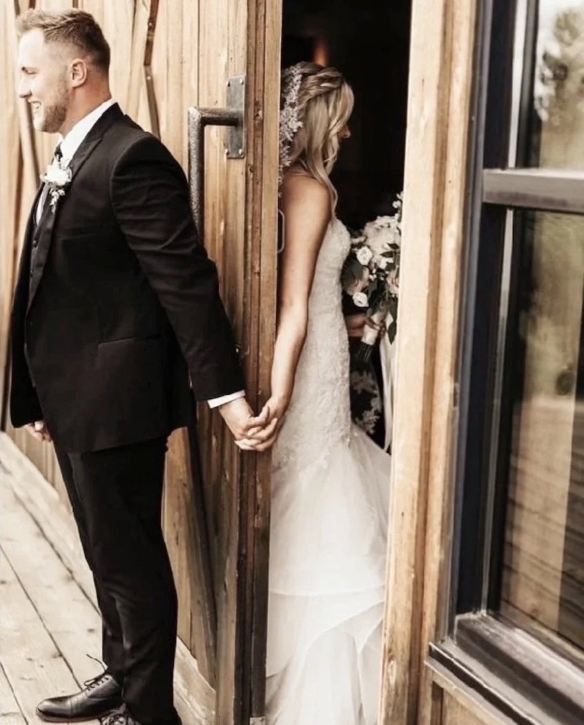 A bride and groom holding hands from back, standing on different sides of a wooden door, with the groom on the left in a black tuxedo and the bride on the right in a wedding gown, with a bouquet, inside a rustic venue.