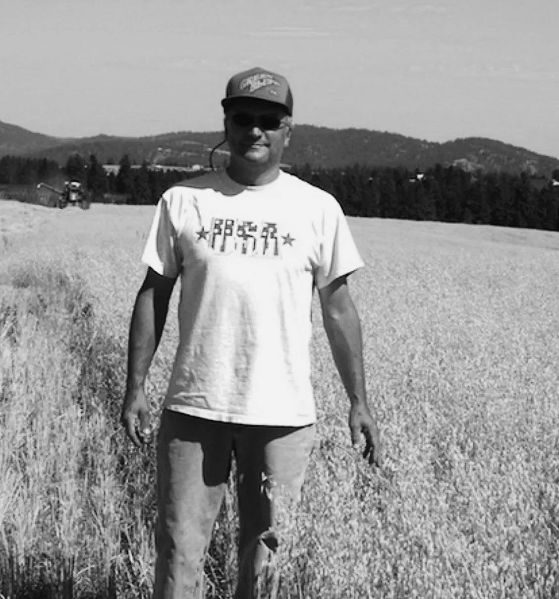 A man wearing sunglasses, a cap, and a T-shirt standing in a field with trees and mountains in the background.