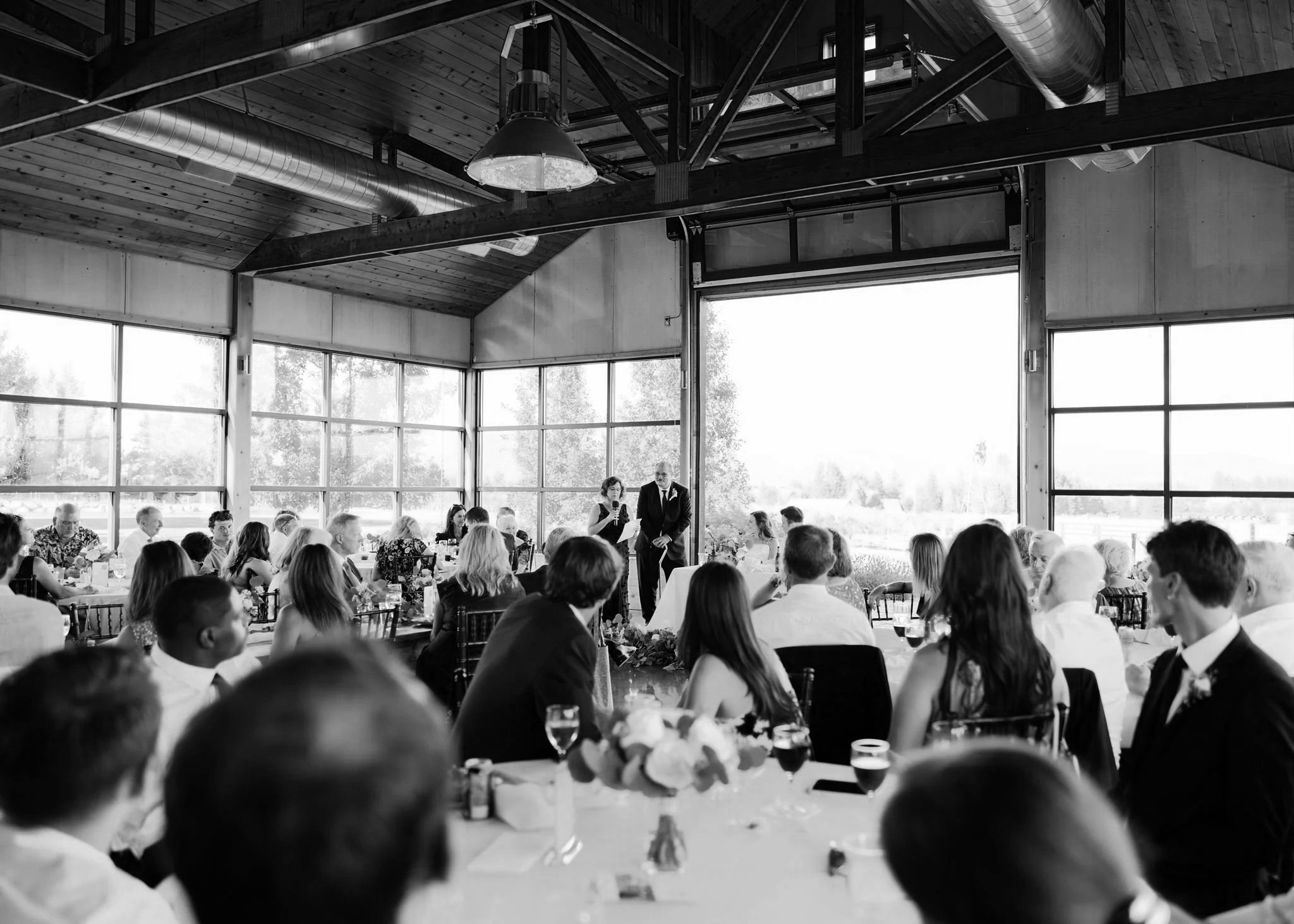 Interior of Trezzi Farm's Barrel Barn with large wrap-around windows, a wooden ceiling, beams, and a long wooden wedding party table set with plates, glasses, and two potted plants at each end, overlooking a scenic landscape of distant mountains.