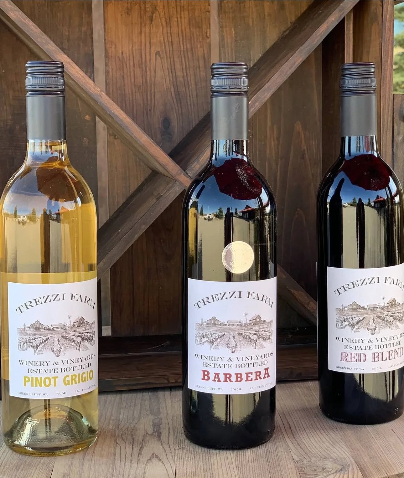 Three bottles of wine from Trezzi Farm Winery & Vineyards on a wooden surface. The left bottle is white wine labeled Pinot Grigio, the middle is red labeled Barbera, and the right is red labeled Red Blend. A wooden wine rack with compartments is in t