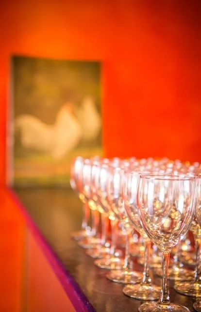 Multiple empty wine glasses arranged in a row on a dark counter, with a blurred painting of swans in the background against a reddish-orange wall.