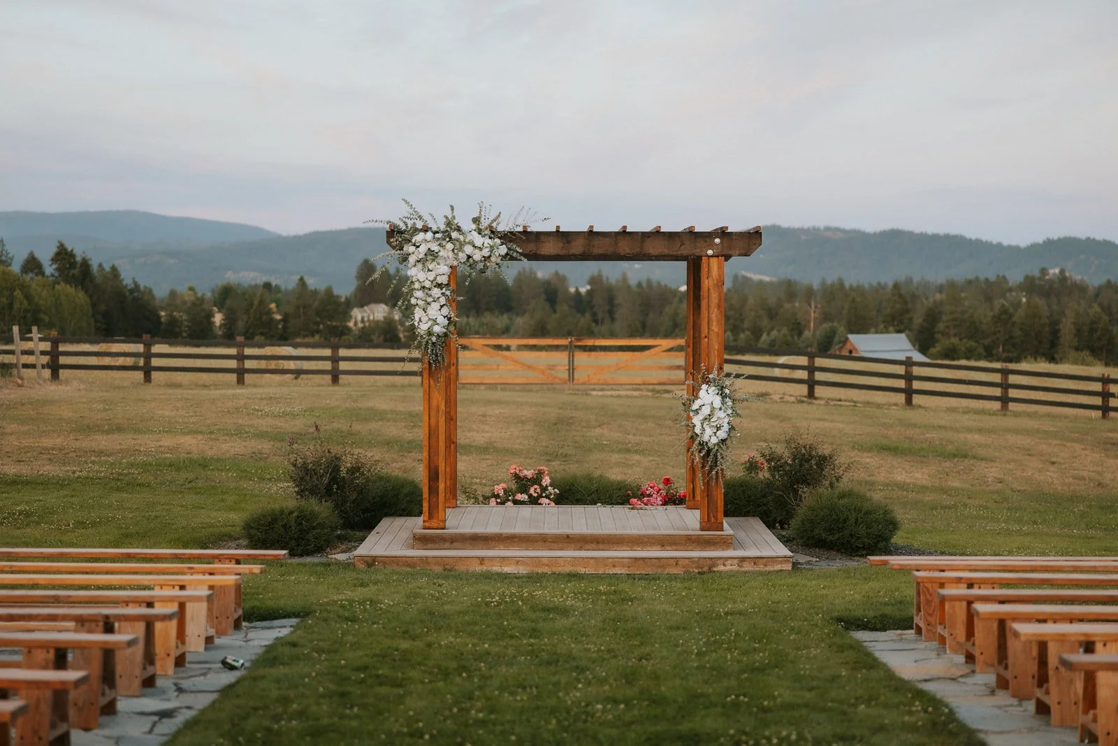 Trezzi Farm's outdoor wedding arbor with a wooden platform decorated with white drapery and greenery, surrounded by benches on a grassy field with a wine barrel and scenic mountain , vineyard, farmland and forest views.