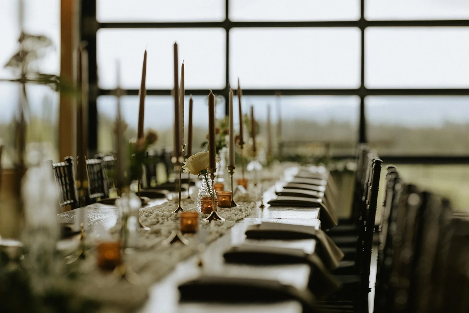 Elegant wedding reception table with taper candles, white roses, amber votives, and place settings by large windows