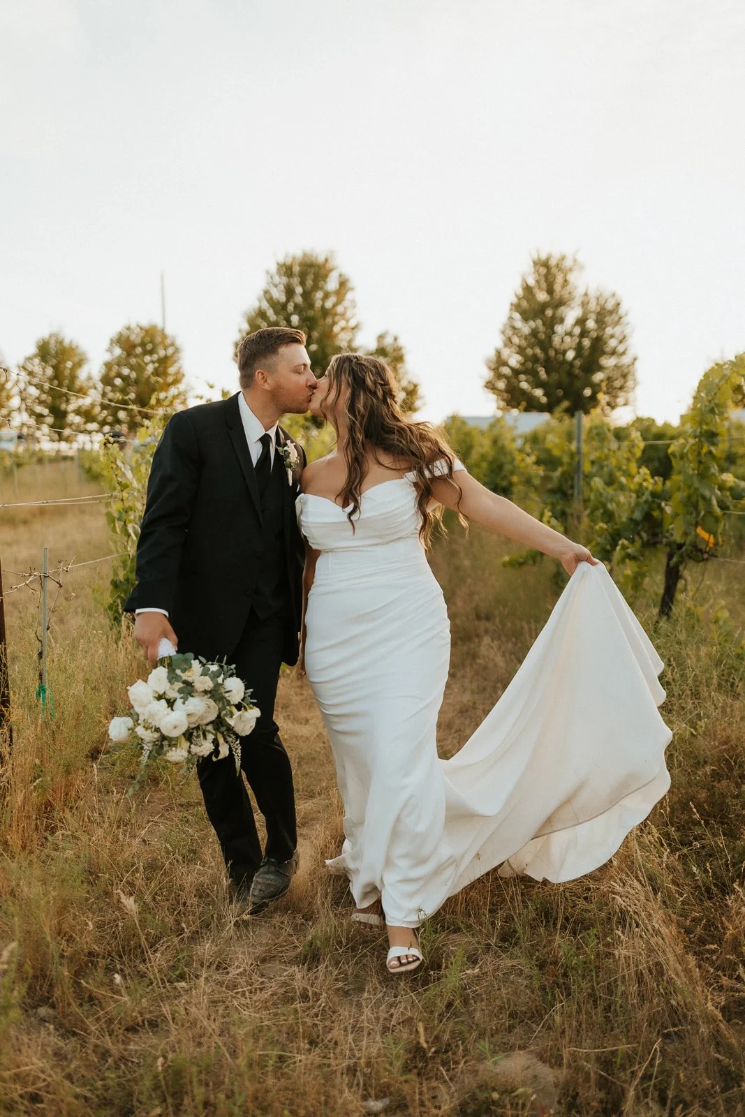 Bride in a white dress and groom in a black suit walking through a vineyard on Green Bluff. with trees in the background Groom is holding brides's bouquet of white flowers.