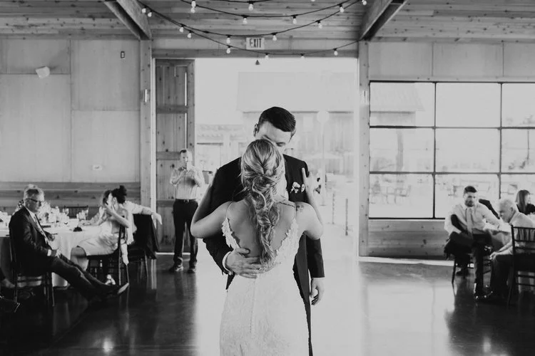 A couple is dancing closely in a rustic and modern barn, with guests seated at tables around them.