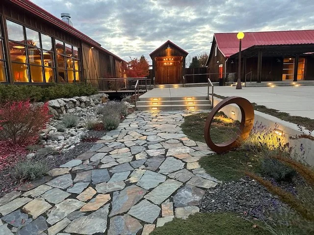 A stone walkway leading to a wooden building with steps and railings, surrounded by landscaped garden and modern outdoor lighting at dusk.