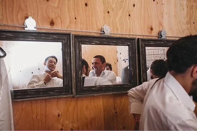 Three groomsmen in white shirts are seen through a set of three framed mirrors on a wooden wall, smiling and interacting with each other.