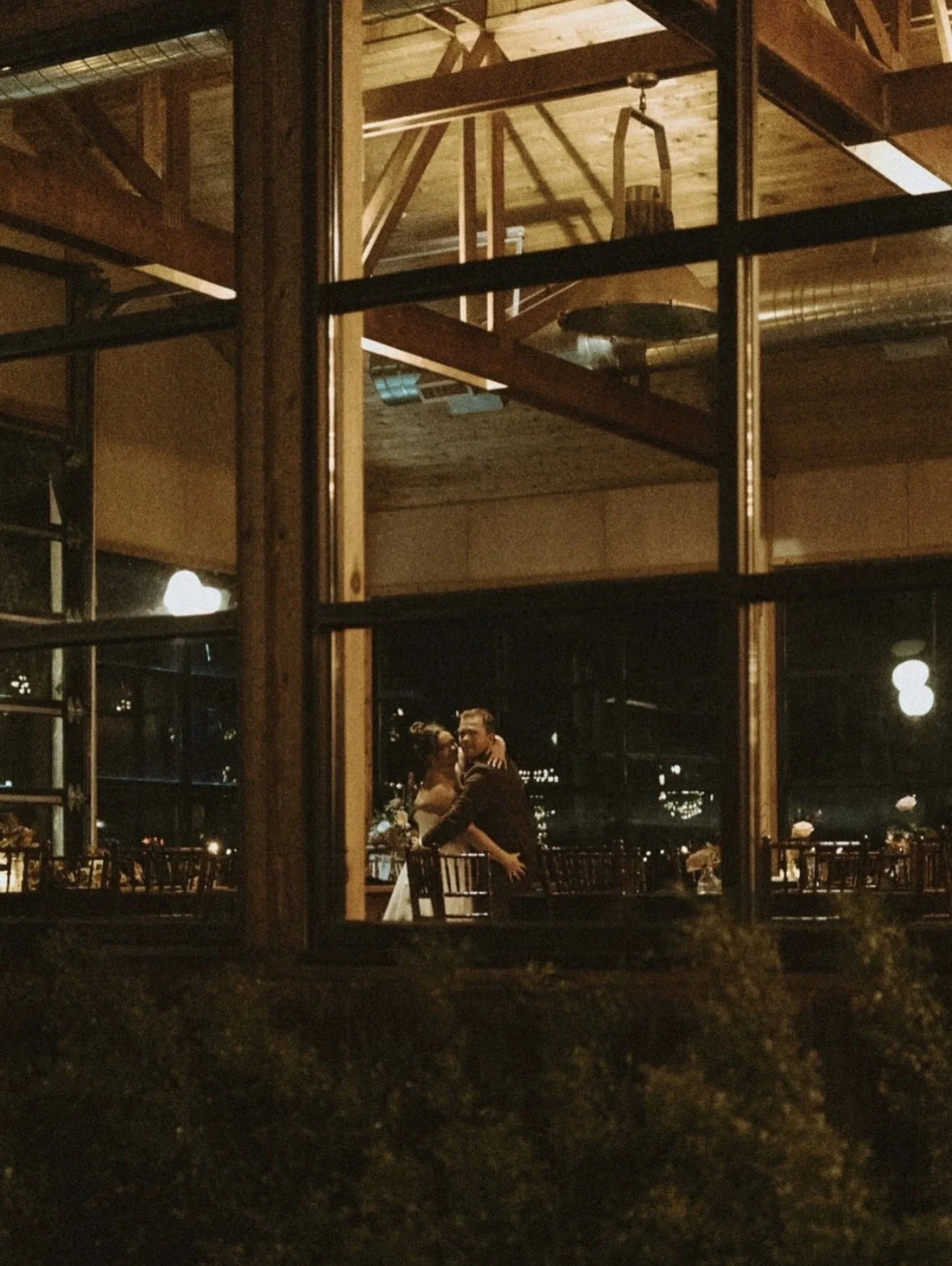 Couple embracing inside a warmly lit restaurant, viewed through large industrial-style windows at night