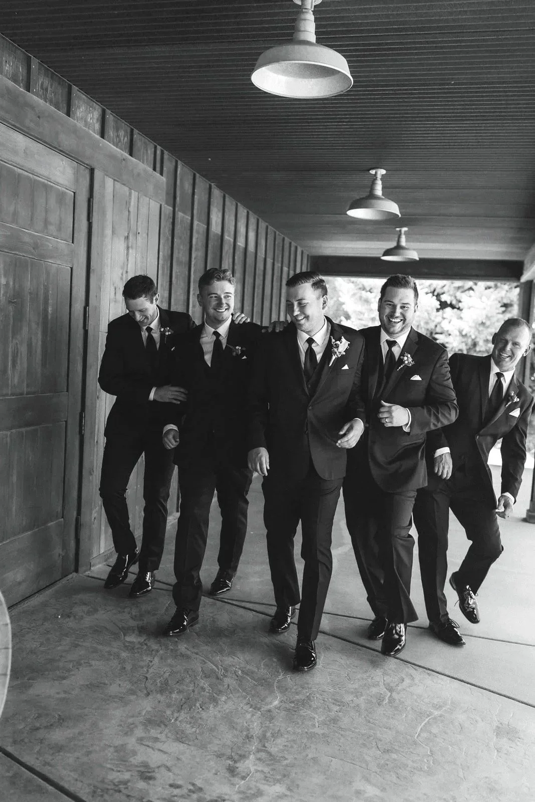 A group of five men in suits walking together with smiles, indoors with wooden walls and ceiling, black and white photo.