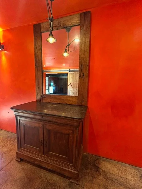 Wooden cabinet with a large mirror attached to the wall, set against an orange painted wall, with hanging lights reflected in the mirror.