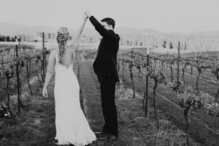 A couple dancing in a vineyard, with the woman in a wedding dress and the man in a suit, holding hands in the air.