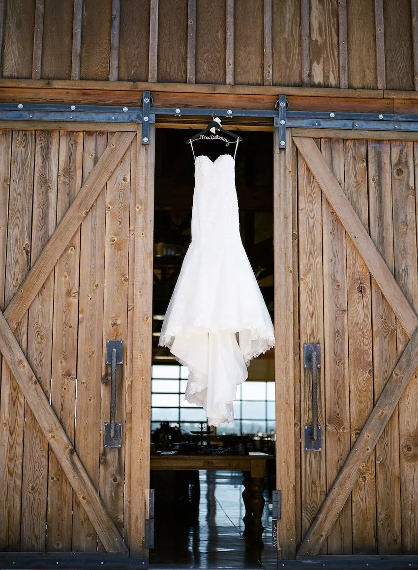 A wedding dress hanging on a hanger in a barn. The barn has wooden sliding doors, open to reveal a rustic interior with tables and windows.