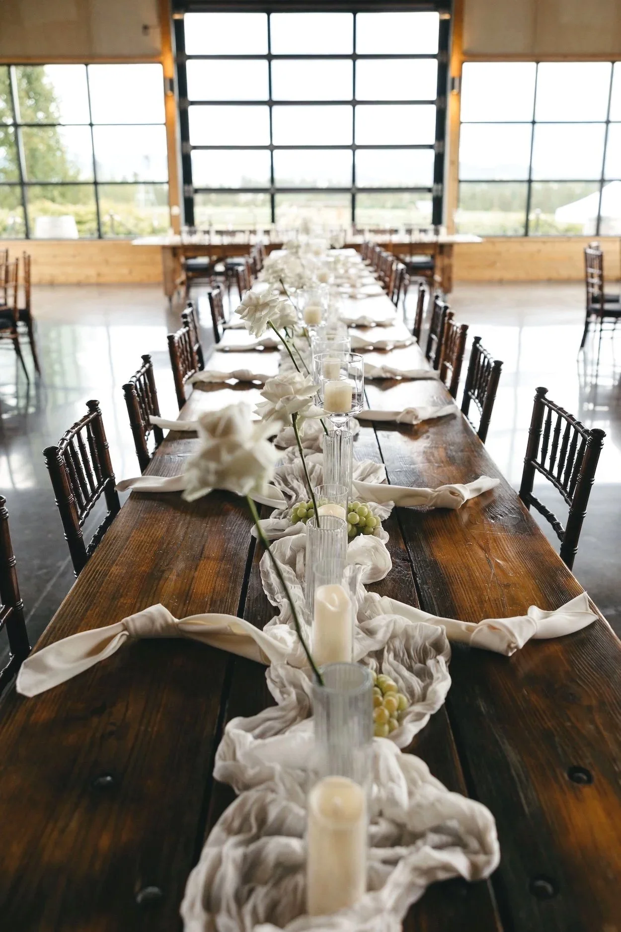 Long rustic wedding reception table with white floral centerpiece, candles, grapes, folded napkins, and dark wood chairs in a sunlit barn venue