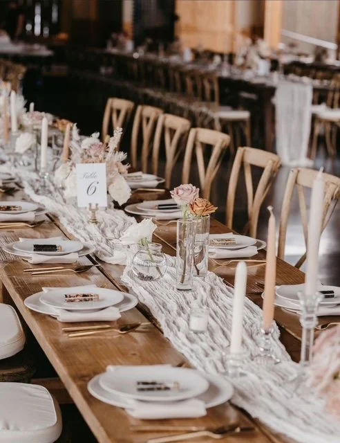 Elegant farm table set with white plates, silverware, and napkins, decorated with vases of flowers and tall candles in a rustic wooden venue.