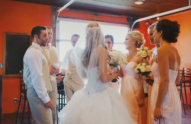 Bride and groom with their wedding party gathering indoors, holding bouquets.