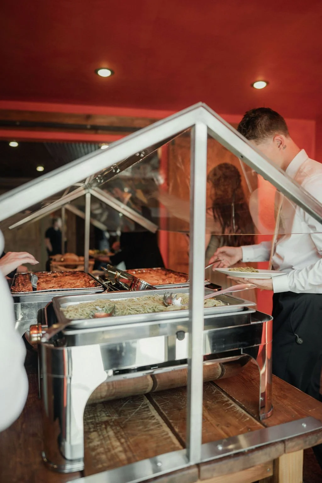 Buffet with trays of pasta and cooked meat, people serving themselves behind a glass shield.