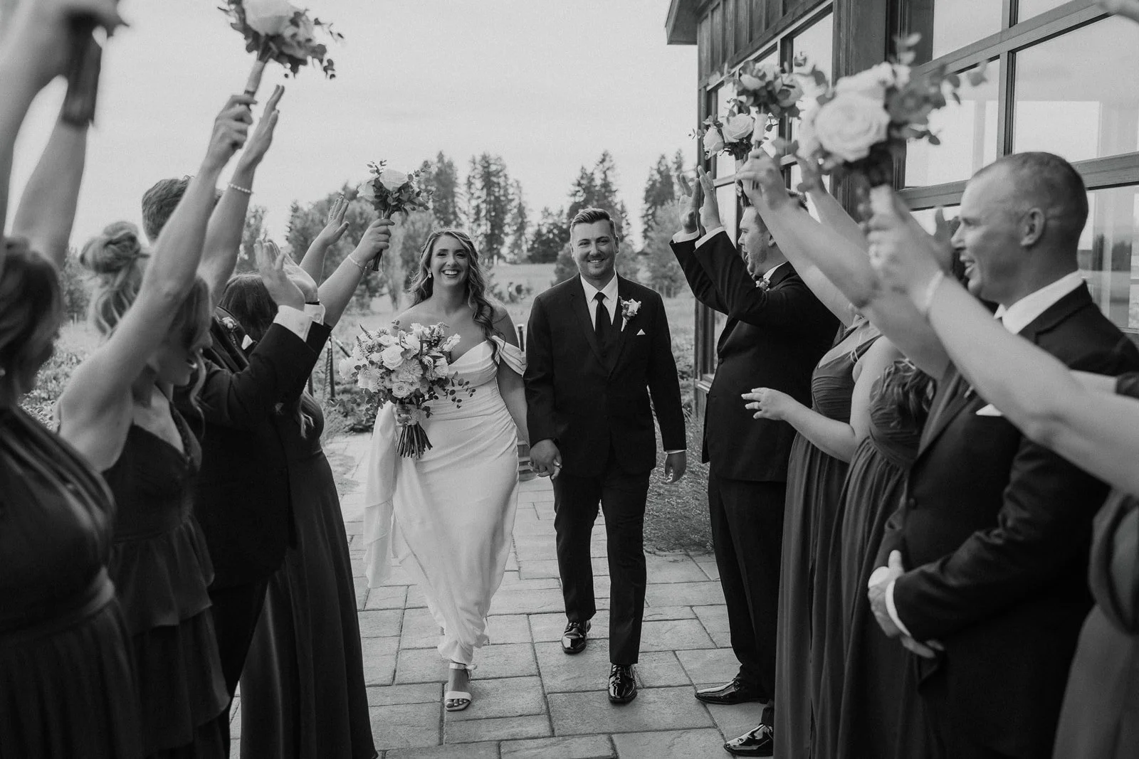 Black and white photo of a bride and groom walking hand in hand through an aisle of wedding guests holding bouquets, outdoors on a veranda with vineyards in the background.