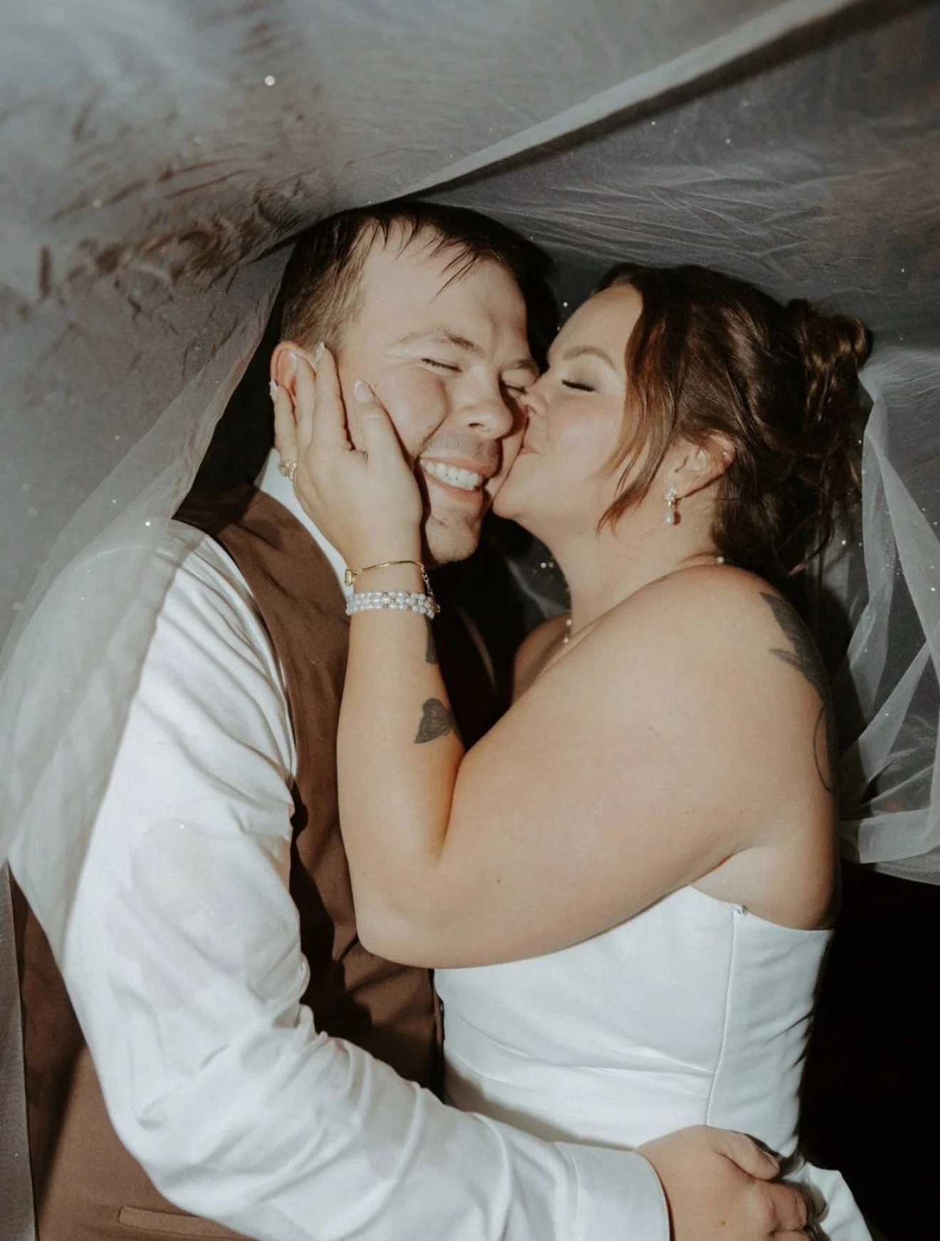 Couple smiling and kissing under a sheer canopy, with the woman wearing a strapless white wedding dress.