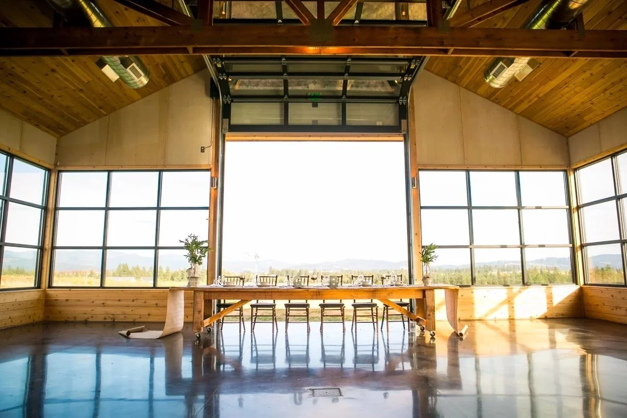 Interior of a bright, rustic modern barn with large windows, wooden ceiling, and a long wooden table set with chairs and tableware, overlooking a scenic landscape.
